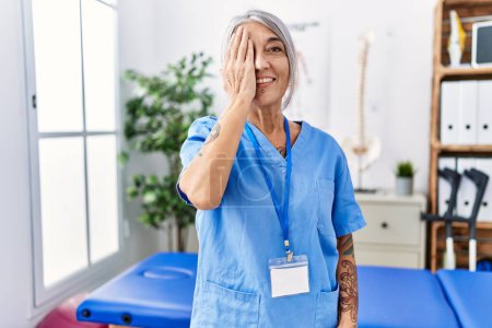 Foto de Mujer de pelo gris de mediana edad que usa uniforme de fisioterapeuta en la clínica médica que cubre un ojo con la mano, sonrisa confiada en la cara y emoción sorpresa. - Imagen libre de derechos
