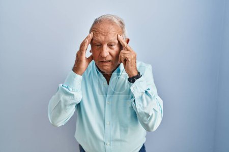 Foto de Hombre mayor con el pelo gris de pie sobre fondo azul con la mano en la cabeza para el dolor en la cabeza porque el estrés. padecer migraña. - Imagen libre de derechos