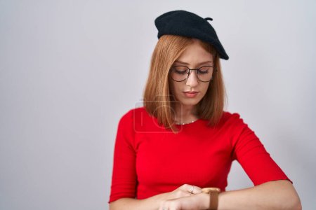 Téléchargez les photos : Young redhead woman standing wearing glasses and beret checking the time on wrist watch, relaxed and confident - en image libre de droit