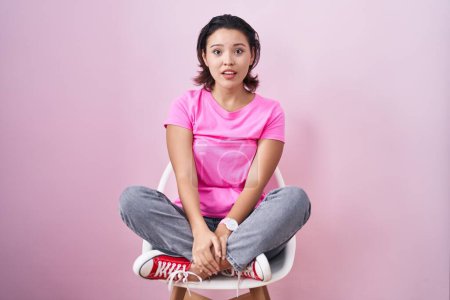 Photo for Hispanic young woman sitting on chair over pink background afraid and shocked with surprise and amazed expression, fear and excited face. - Royalty Free Image