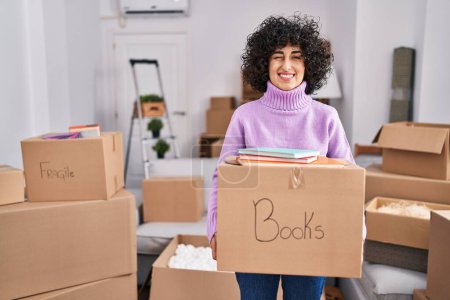 Foto de Young brunette woman with curly hair moving to a new home holding cardboard box winking looking at the camera with sexy expression, cheerful and happy face. - Imagen libre de derechos