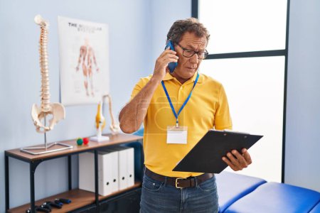 Foto de Fisioterapeuta hombre de mediana edad hablando en el teléfono inteligente en la clínica de fisioterapia - Imagen libre de derechos
