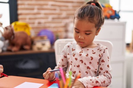 Téléchargez les photos : Adorable étudiante hispanique assise sur la table dessinant sur papier à la maternelle - en image libre de droit