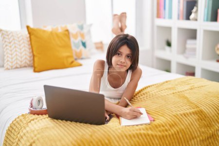 Photo for Adorable hispanic girl using laptop writing on notebook at bedroom - Royalty Free Image