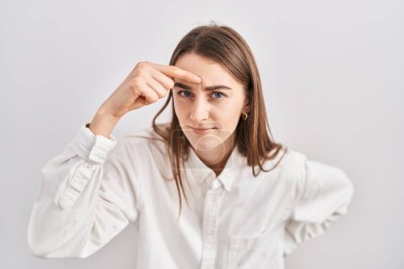 Foto de Young caucasian woman standing over isolated background pointing unhappy to pimple on forehead, ugly infection of blackhead. acne and skin problem - Imagen libre de derechos