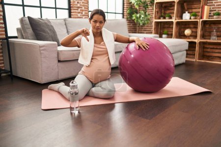 Photo pour Jeune femme enceinte assise sur un tapis de yoga avec balle pilates avec visage en colère, signe négatif montrant aversion avec les pouces vers le bas, concept de rejet - image libre de droit