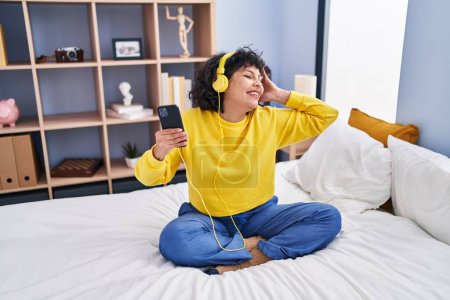 Téléchargez les photos : Jeune belle femme hispanique dansant et écoutant de la musique assise sur le lit dans la chambre - en image libre de droit