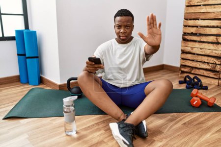 Téléchargez les photos : Young african man sitting on training mat at the gym using smartphone doing stop sing with palm of the hand. warning expression with negative and serious gesture on the face. - en image libre de droit