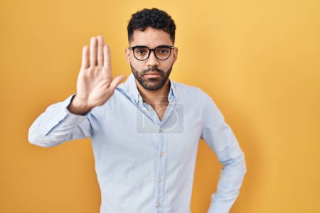 Photo for Hispanic man with beard standing over yellow background doing stop sing with palm of the hand. warning expression with negative and serious gesture on the face. - Royalty Free Image