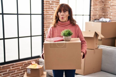 Photo for Middle age hispanic woman moving to a new home holding cardboard box making fish face with mouth and squinting eyes, crazy and comical. - Royalty Free Image