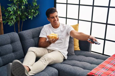 Téléchargez les photos : Young hispanic man watching movie eating chips potatoes at home - en image libre de droit