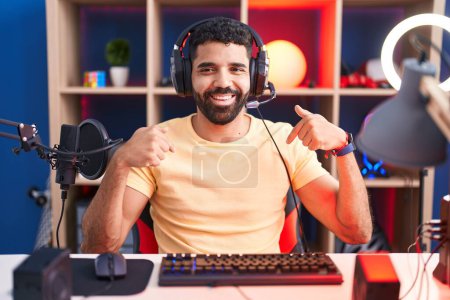Foto de Hombre hispano con barba jugando videojuegos con auriculares que se ven confiados con sonrisa en la cara, señalándose con los dedos orgullosos y felices. - Imagen libre de derechos