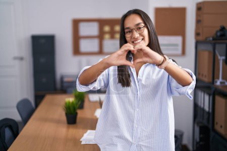 Téléchargez les photos : Jeune femme hispanique au bureau souriant d'amour faisant forme de symbole de coeur avec les mains. concept romantique. - en image libre de droit