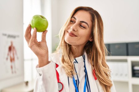 Téléchargez les photos : Jeune femme blonde portant l'uniforme de médecin tenant pomme à la clinique - en image libre de droit