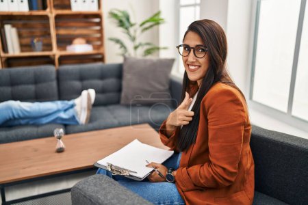 Foto de Young hispanic woman working as psychology counselor cheerful with a smile on face pointing with hand and finger up to the side with happy and natural expression - Imagen libre de derechos