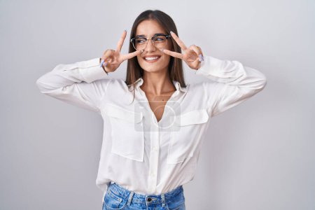 Téléchargez les photos : Jeune femme brune portant des lunettes faisant symbole de paix avec les doigts sur le visage, souriant gai montrant la victoire - en image libre de droit