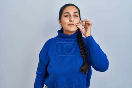 Foto de Young hispanic woman standing over isolated background mouth and lips shut as zip with fingers. secret and silent, taboo talking - Imagen libre de derechos