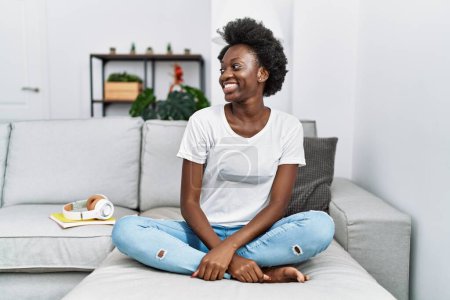 Téléchargez les photos : African young woman sitting on the sofa at home looking away to side with smile on face, natural expression. laughing confident. - en image libre de droit