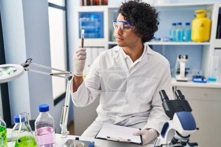 Photo for Young hispanic man wearing scientist uniform holding clipboard and test tube at laboratory - Royalty Free Image