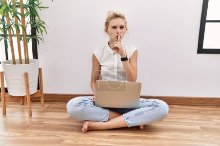 Photo for Young blonde woman using computer laptop sitting on the floor at the living room asking to be quiet with finger on lips. silence and secret concept. - Royalty Free Image