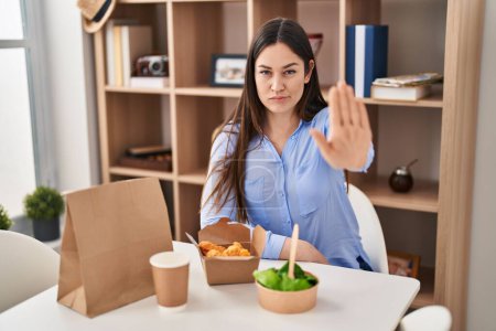 Téléchargez les photos : Jeune femme brune manger emporter de la nourriture à la maison avec la main ouverte faisant stop signe avec expression sérieuse et confiante, geste de défense - en image libre de droit
