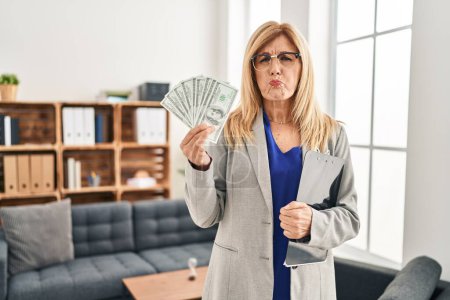 Foto de Middle age blonde woman working at therapy consultation office holding money puffing cheeks with funny face. mouth inflated with air, catching air. - Imagen libre de derechos