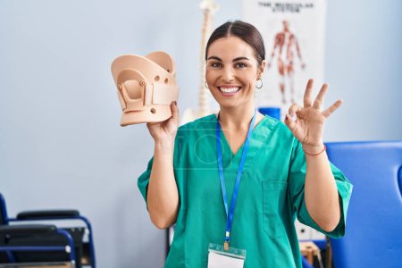 Photo for Young hispanic woman holding cervical neck collar at the clinic doing ok sign with fingers, smiling friendly gesturing excellent symbol - Royalty Free Image