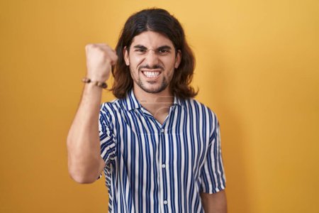 Photo for Hispanic man with long hair standing over yellow background angry and mad raising fist frustrated and furious while shouting with anger. rage and aggressive concept. - Royalty Free Image