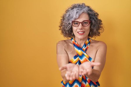 Foto de Mujer de mediana edad con el pelo gris de pie sobre fondo amarillo sonriendo con las palmas de las manos juntas recibiendo o dando gesto. retención y protección - Imagen libre de derechos