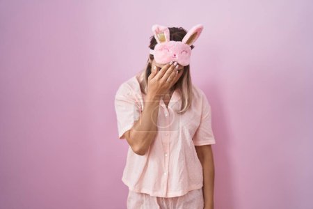 Foto de Mujer rubia caucásica con máscara para dormir y pijama con expresión triste cubriendo la cara con las manos mientras llora. concepto de depresión. - Imagen libre de derechos