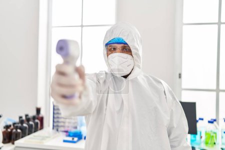 Téléchargez les photos : Jeune homme latino-scientifique portant un thermomètre de maintien uniforme de protection covide au laboratoire - en image libre de droit