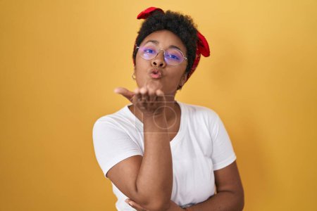Téléchargez les photos : Jeune femme afro-américaine debout sur fond jaune regardant la caméra souffler un baiser avec la main sur l'air étant belle et sexy. expression d'amour. - en image libre de droit