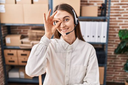 Foto de Mujer caucásica joven que trabaja en el comercio electrónico de pequeñas empresas con auriculares sonriendo feliz haciendo signo de ok con la mano en el ojo mirando a través de los dedos - Imagen libre de derechos