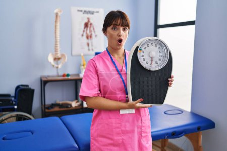 Photo for Young brunette woman as nutritionist holding weighing machine scared and amazed with open mouth for surprise, disbelief face - Royalty Free Image