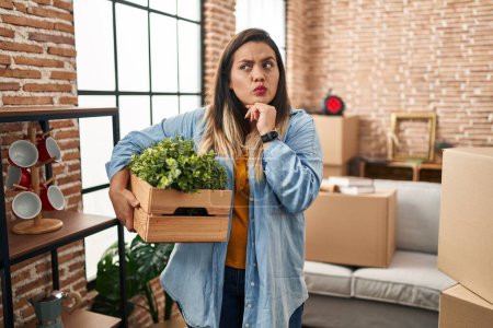 Téléchargez les photos : Young hispanic woman moving to a new home holding plants serious face thinking about question with hand on chin, thoughtful about confusing idea - en image libre de droit