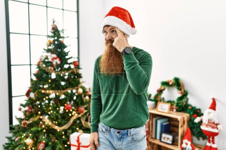 Téléchargez les photos : Homme rousse à longue barbe portant un chapeau de Noël par l'arbre de Noël pointant malheureux de boutons sur le front, infection laid de la tête noire. acné et problème de peau - en image libre de droit