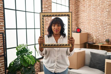 Photo for Hispanic woman at new home holding empty frame relaxed with serious expression on face. simple and natural looking at the camera. - Royalty Free Image