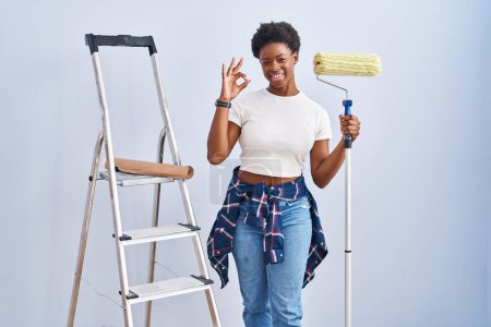 Foto de Mujer afroamericana sosteniendo rodillo pintor sonriendo positiva haciendo signo ok con la mano y los dedos. expresión exitosa. - Imagen libre de derechos