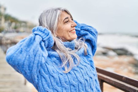 Téléchargez les photos : Femme aux cheveux gris d'âge moyen souriant confiante détendue avec les mains sur la tête au bord de la mer - en image libre de droit