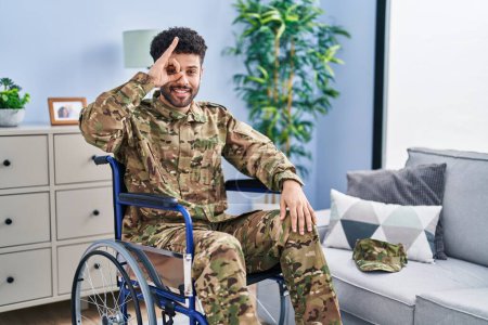 Foto de Hombre árabe vistiendo uniforme camuflaje ejército sentado en silla de ruedas haciendo buen gesto con la mano sonriente, los ojos mirando a través de los dedos con cara feliz. - Imagen libre de derechos