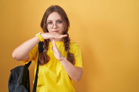 Photo for Young caucasian woman wearing student backpack over yellow background doing time out gesture with hands, frustrated and serious face - Royalty Free Image