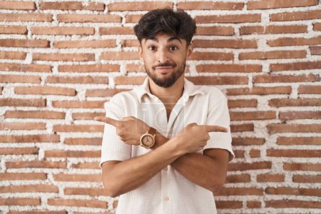 Foto de Arab man with beard standing over bricks wall background pointing to both sides with fingers, different direction disagree - Imagen libre de derechos