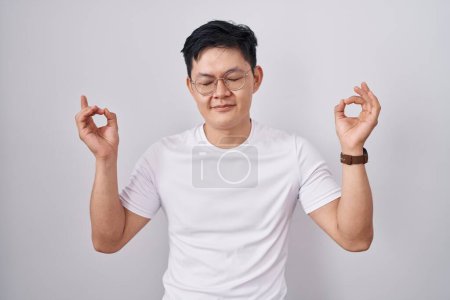 Photo for Young asian man standing over white background relaxed and smiling with eyes closed doing meditation gesture with fingers. yoga concept. - Royalty Free Image