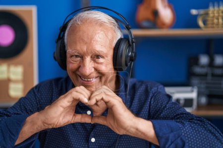 Téléchargez les photos : Senior homme aux cheveux gris artiste écoutant de la musique faire un geste de cœur avec les mains au studio de musique - en image libre de droit