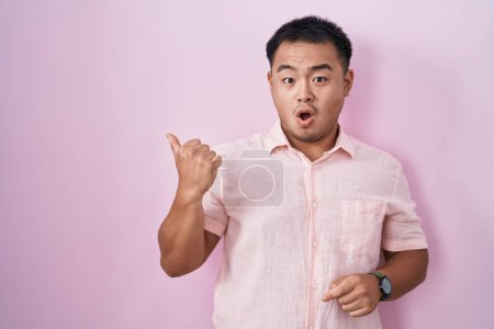 Foto de Chinese young man standing over pink background surprised pointing with hand finger to the side, open mouth amazed expression. - Imagen libre de derechos