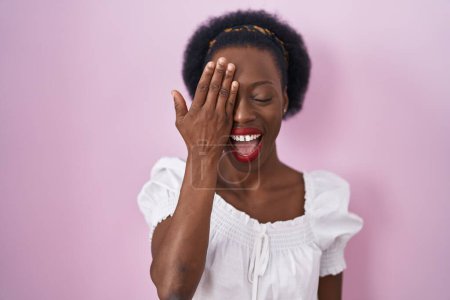 Foto de Mujer africana con el pelo rizado de pie sobre fondo rosa que cubre un ojo con la mano, sonrisa confiada en la cara y emoción sorpresa. - Imagen libre de derechos