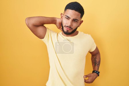 Photo for Young hispanic man standing over yellow background suffering of neck ache injury, touching neck with hand, muscular pain - Royalty Free Image