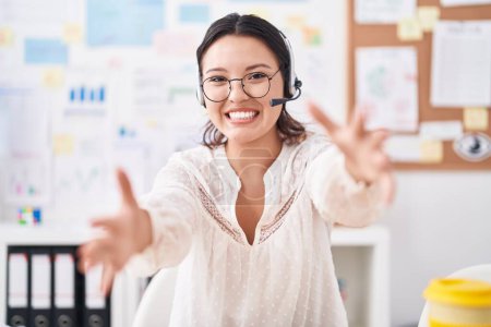Téléchargez les photos : Hispanic young woman working at the office wearing headset and glasses looking at the camera smiling with open arms for hug. cheerful expression embracing happiness. - en image libre de droit