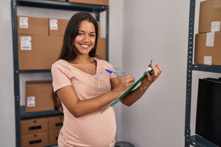 Photo for Young latin woman pregnant business worker writing on clipboard at office - Royalty Free Image