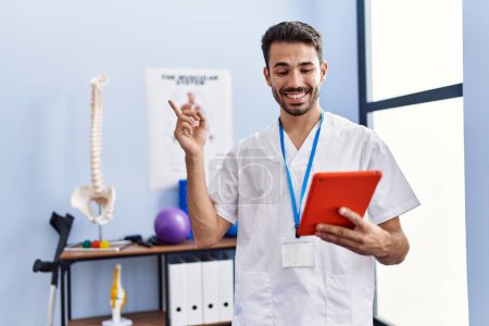Photo for Young hispanic physiotherapist man doing online appointment smiling happy pointing with hand and finger to the side - Royalty Free Image